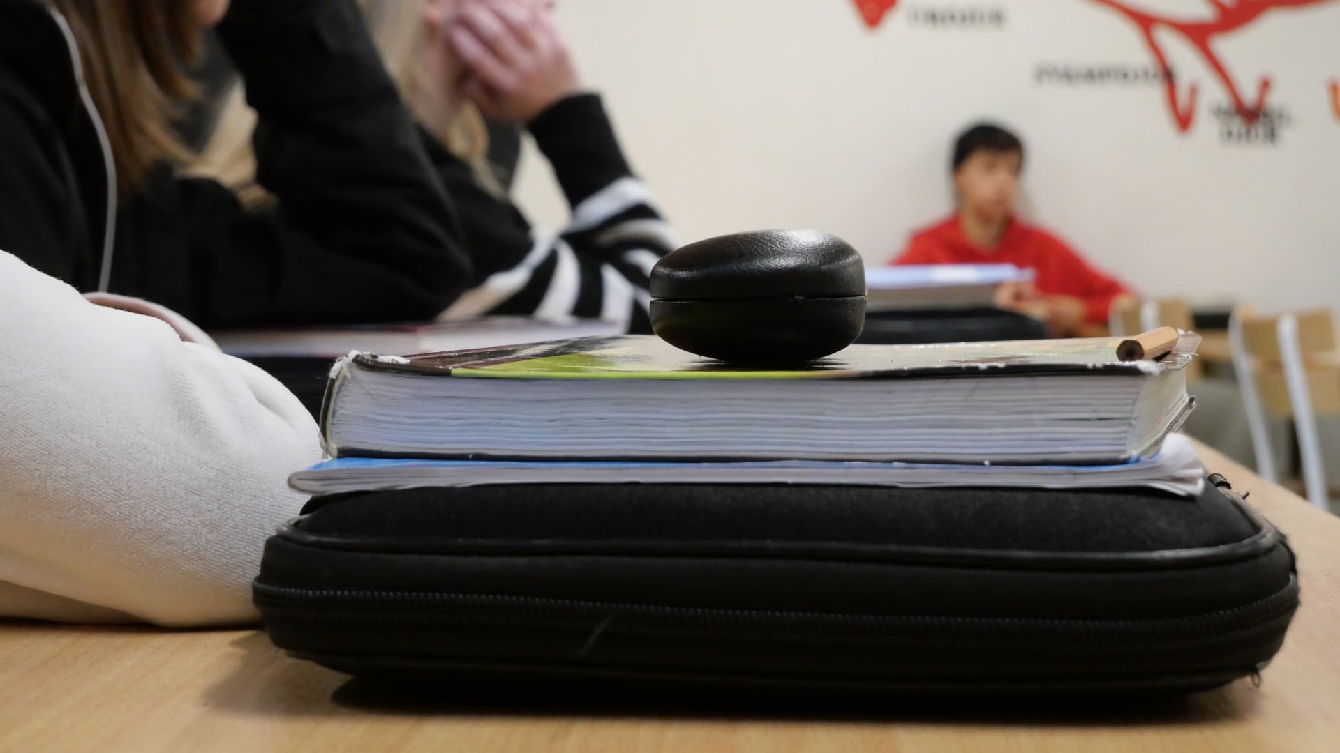 Bilden visar en bok och ett datorfodral som ligger på en skilbänk. I bakgrunden sitter elever i ett klassrum